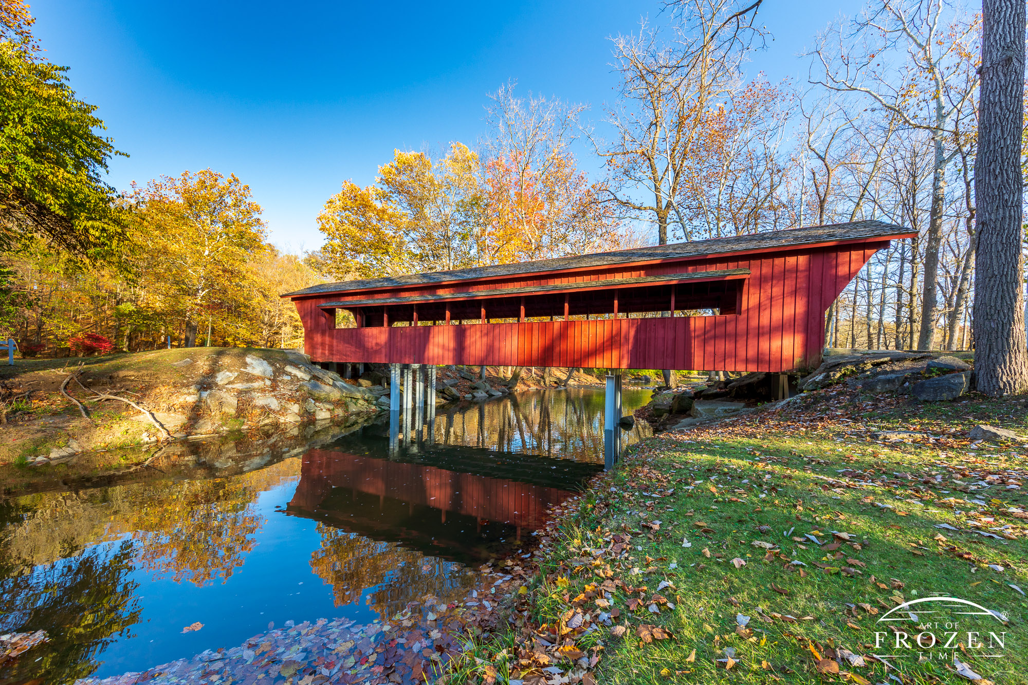 The Ross Covered Bridge resides in Tawawa Park, Sidney Ohio.  The wood bridge (also known as Sidney Ohio’s covered bridge) was constructed in 1971 but drew its design from central Ohio pioneer bridge builder Rueben L. Partridge.  Today, the bridge spans Tawawa Creek as it meanders through the park.  In this golden hour image, the red bridge basks in warm light and the thinning autumn leaves and clear blue sky.