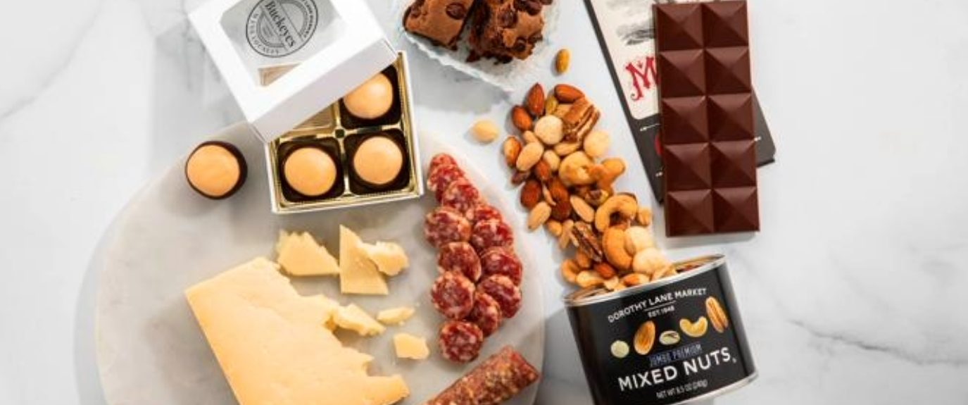 Assortment of gourmet gifts from Dorothy Lane Market including chocolates, nuts, cheese, sausage, brownies, and gift boxes displayed on a marble surface.