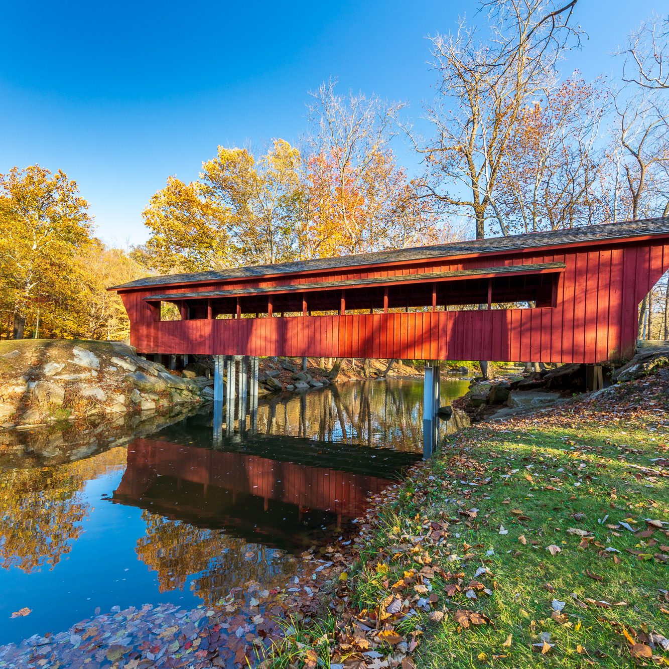 The Ross Covered Bridge resides in Tawawa Park, Sidney Ohio.  The wood bridge (also known as Sidney Ohio’s covered bridge) was constructed in 1971 but drew its design from central Ohio pioneer bridge builder Rueben L. Partridge.  Today, the bridge spans Tawawa Creek as it meanders through the park.  In this golden hour image, the red bridge basks in warm light and the thinning autumn leaves and clear blue sky.